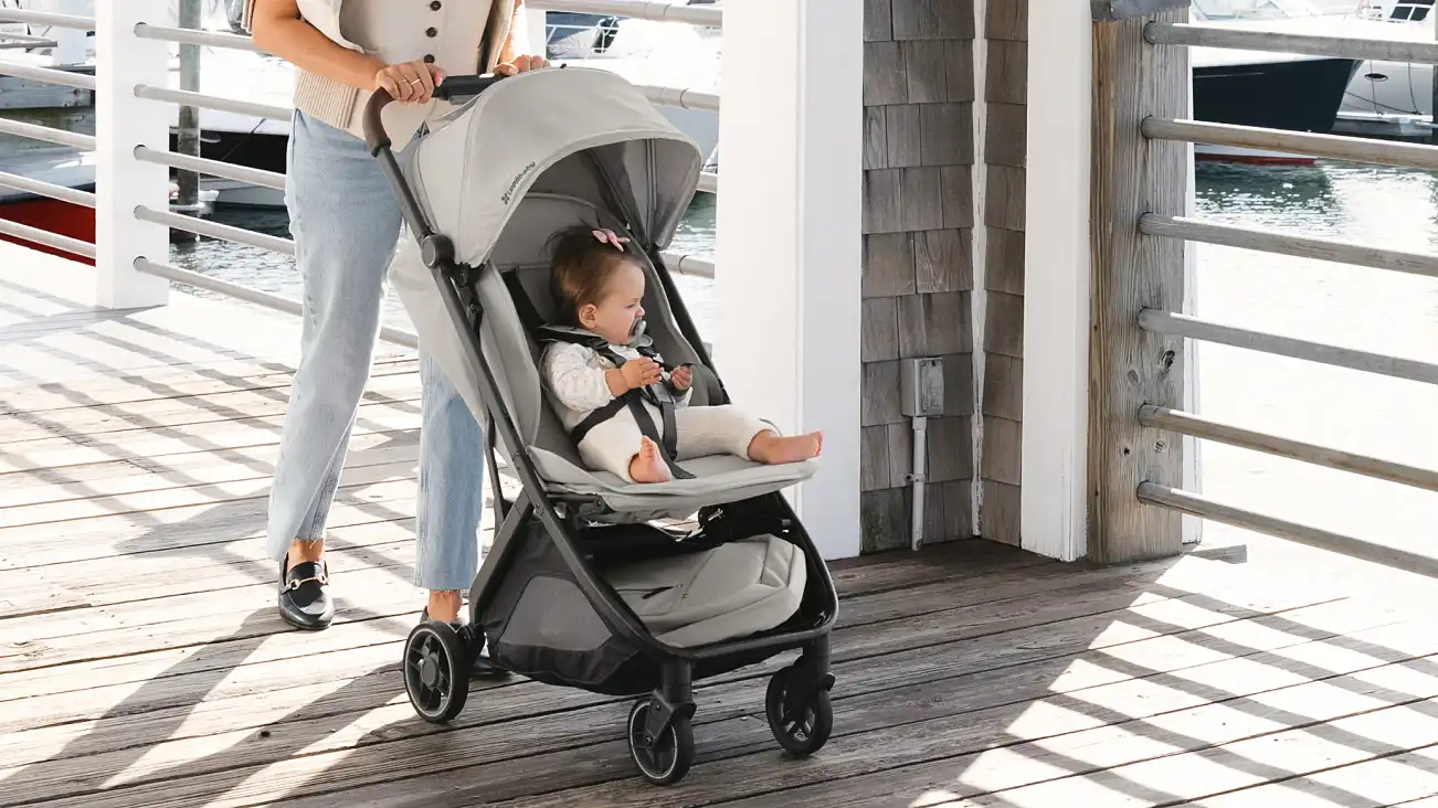 Child harnessed in a Minu V3 with the leg rest raised, pushed by an adult along a marina walkway